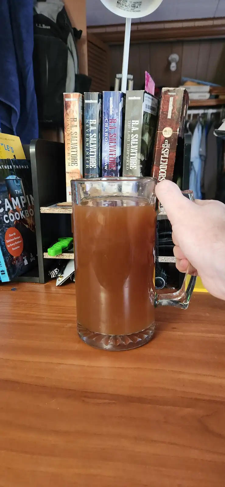 A hand holds a large glass mug filled with a brown liquid, likely beer, on a wooden surface. Behind it, a bookshelf displays…