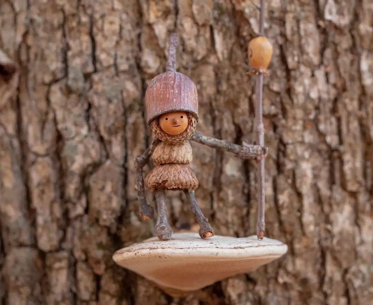 A whimsical, handcrafted figurine made from acorns and twigs stands on a mushroom attached to a tree trunk, holding a stick…