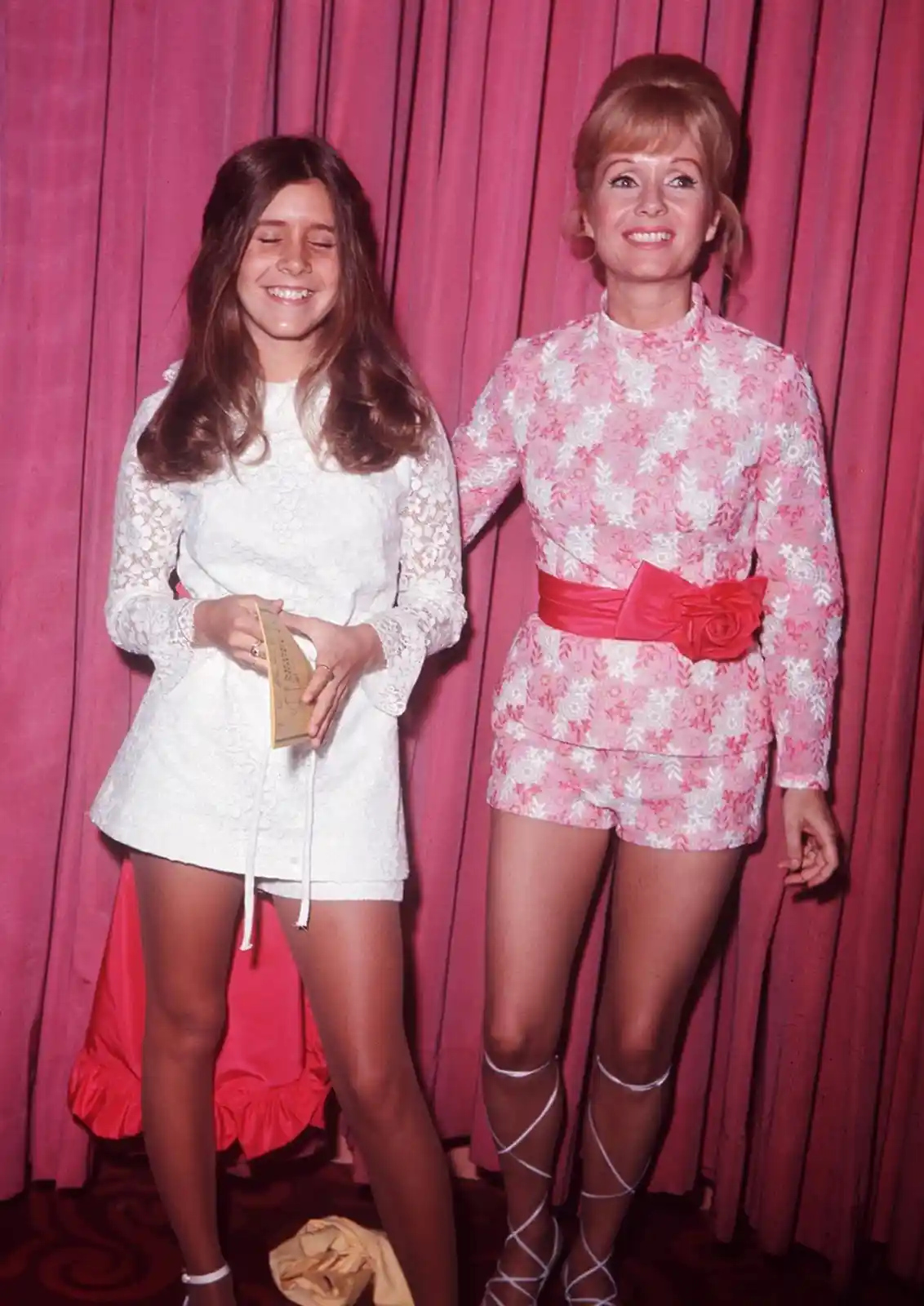 Two women pose in front of a pink curtain; the woman on the left wears a white lace dress and smiles with eyes closed, while…