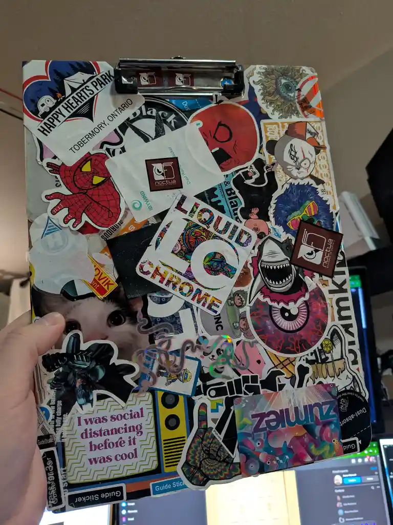 A hand holds a clipboard completely covered in colorful, overlapping stickers featuring cartoon characters, logos, and…