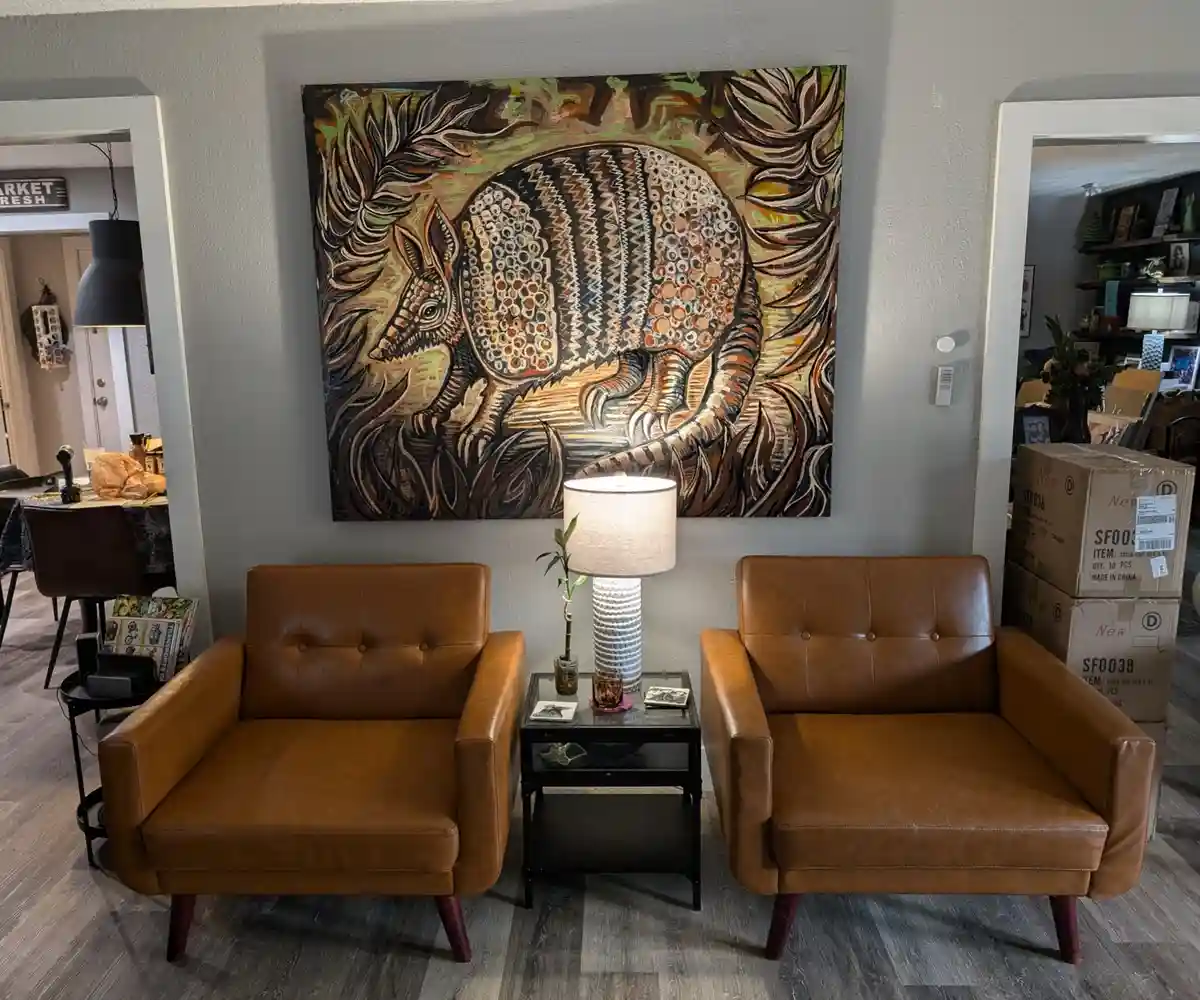 A living room features two brown leather armchairs flanking a small black side table with a lamp and plant. Above them hangs…