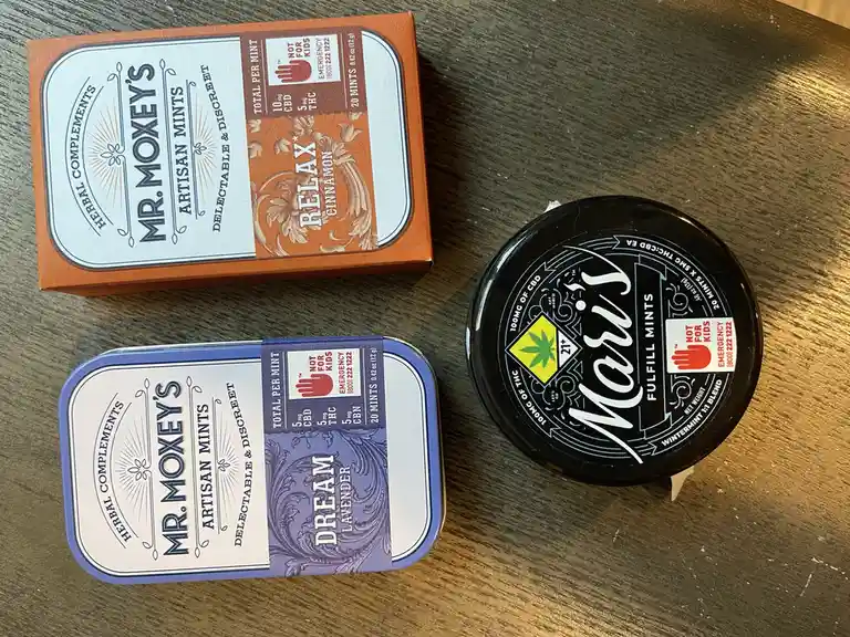 Three cannabis-infused mint containers are arranged on a wooden surface: two Mr. Moxey's tins labeled 'Relax Cinnamon' and…