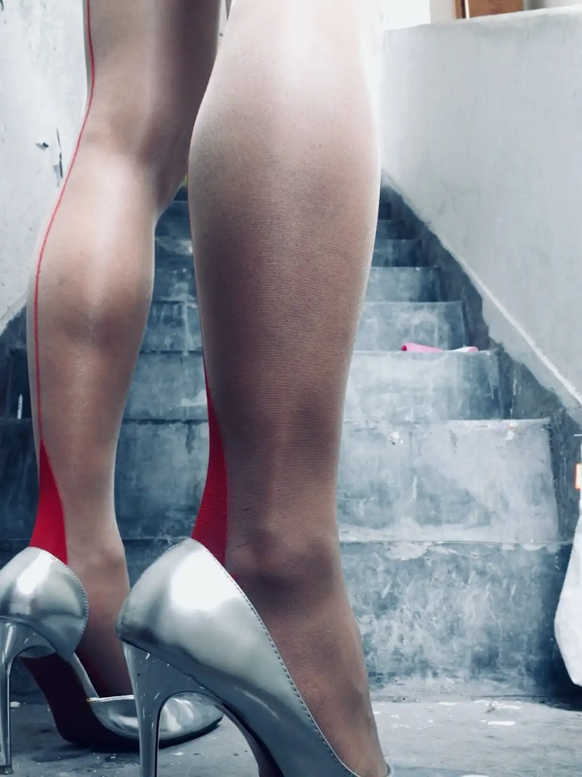 A close-up shot shows a person's legs wearing sheer black stockings with a visible red garter strap running up the left…