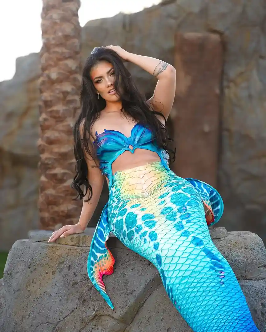 A woman with long dark hair poses confidently while wearing a colorful, scale-patterned mermaid tail costume with a blue and…