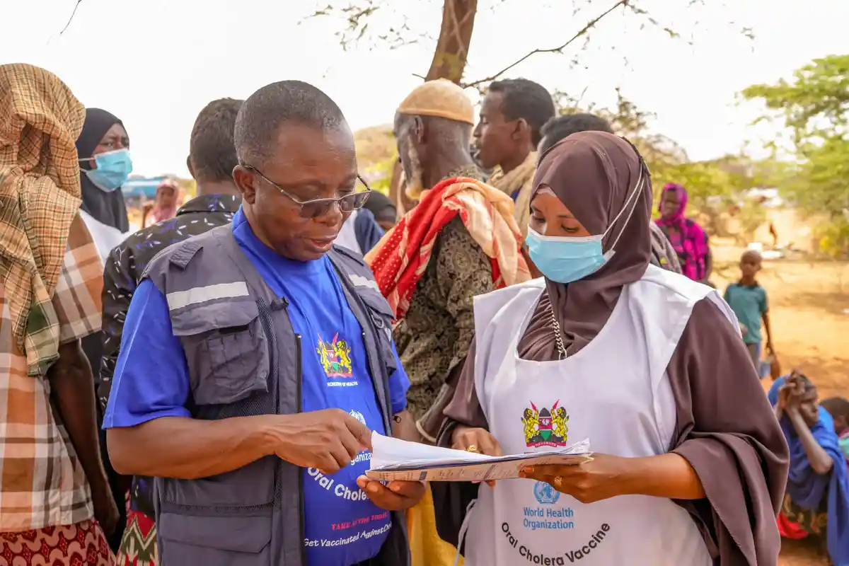 Two health workers in field vests with 'World Health Organization' and 'Ministry of Health' logos are reviewing documents…