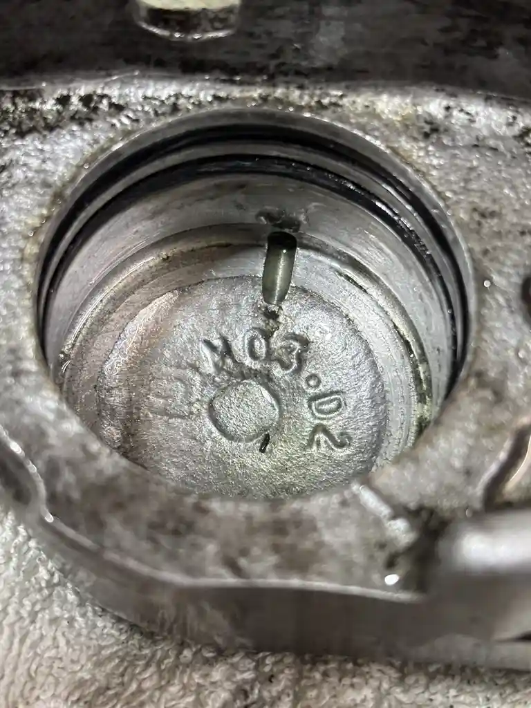 A close-up photo of a worn, metallic mechanical part, possibly a valve or fitting, showing an embossed marking '203.02' on…