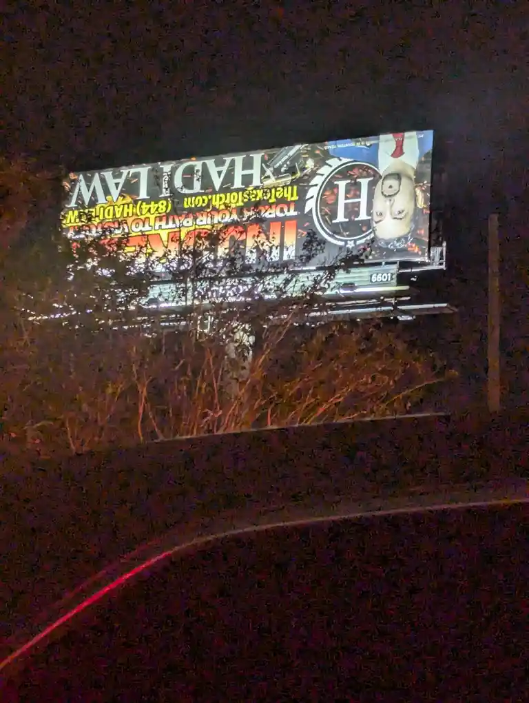 A nighttime photo of a large, illuminated billboard for 'Hadi Law' featuring a man's face upside down, with the tagline…