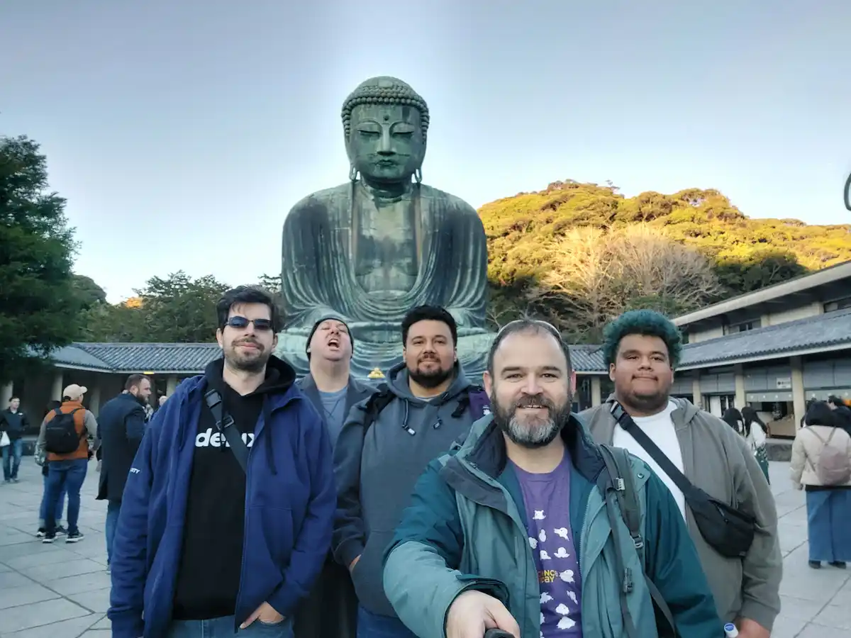 Five men pose for a selfie in front of the giant bronze Great Buddha statue at Kamakura, Japan. One man in the center is…