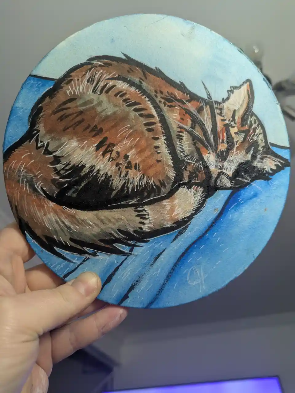 A hand holds up a circular watercolor painting of a brown and black cat curled up sleeping on a blue background. The cat is…