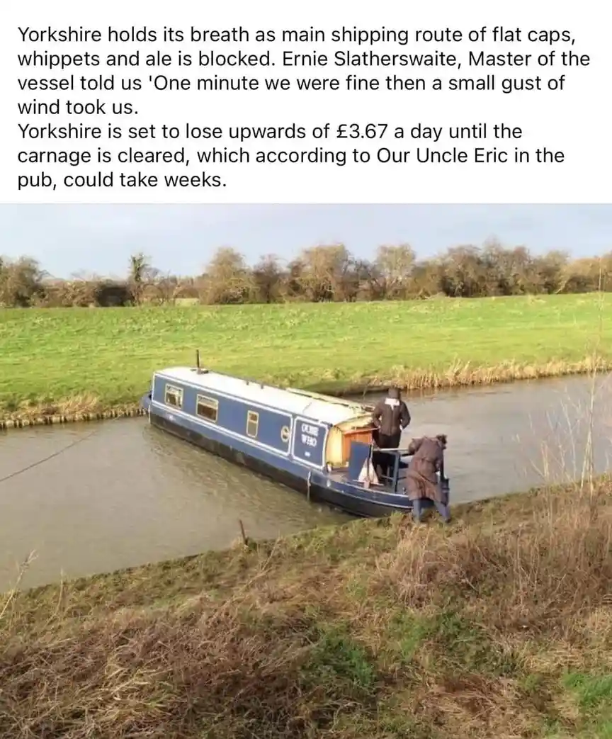A blue and white narrowboat is stuck sideways in a canal, partially on the grassy bank. Two men in coats are on the boat and…