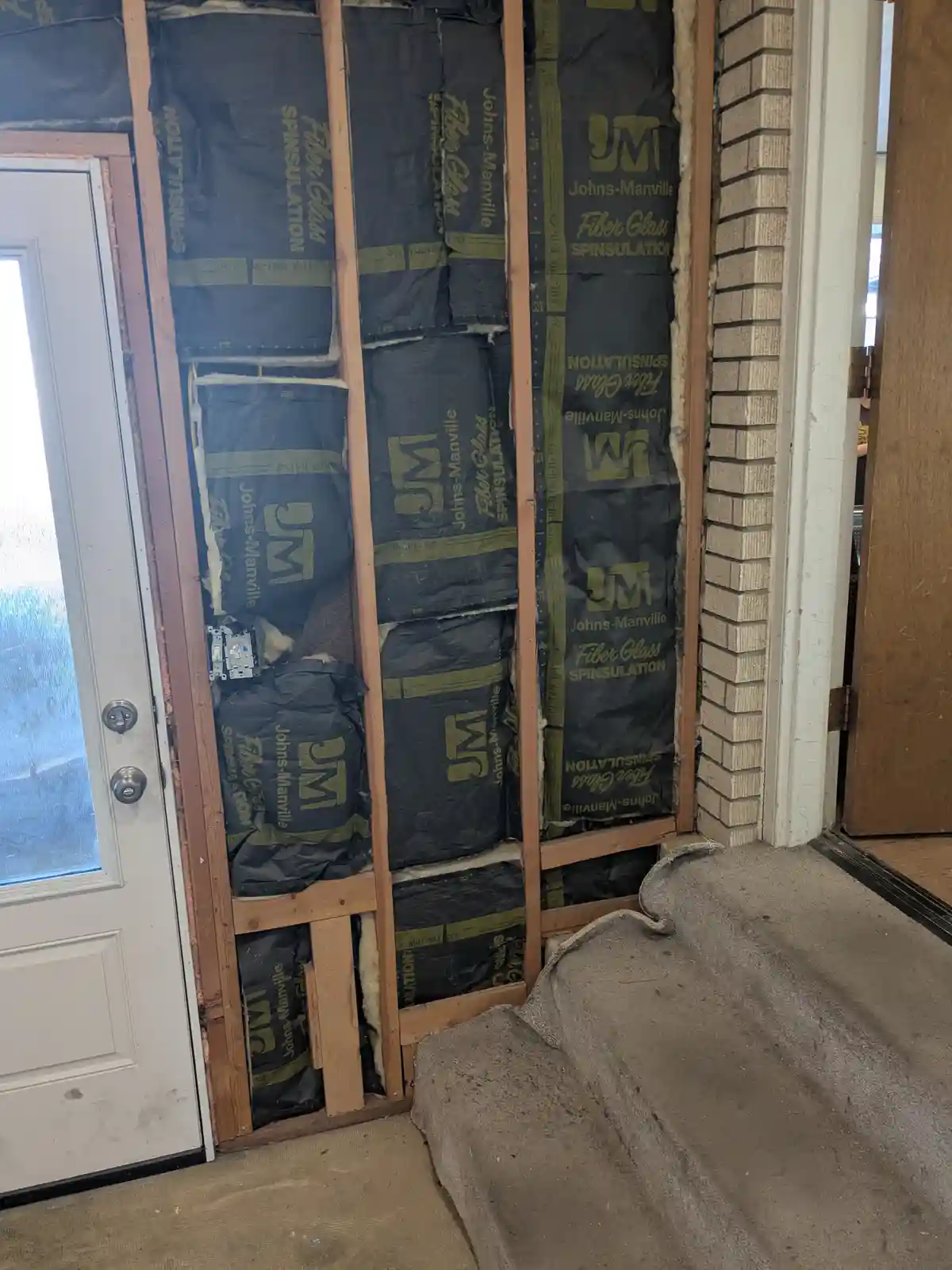 A photo shows a doorway under renovation, with exposed wooden framing filled with black Johns-Manville fiberglass insulation…