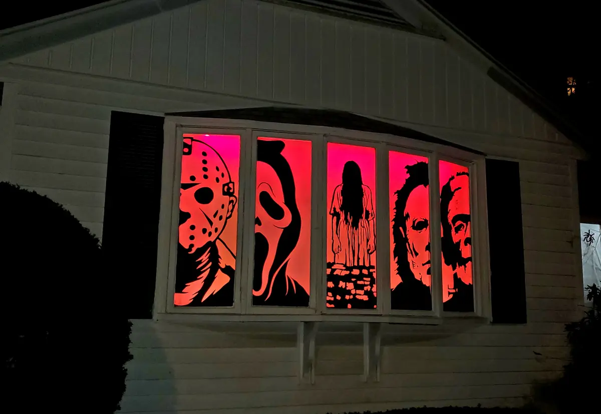 A nighttime photo of a white house's bay window, where each pane is decorated with a black silhouette of a horror movie…