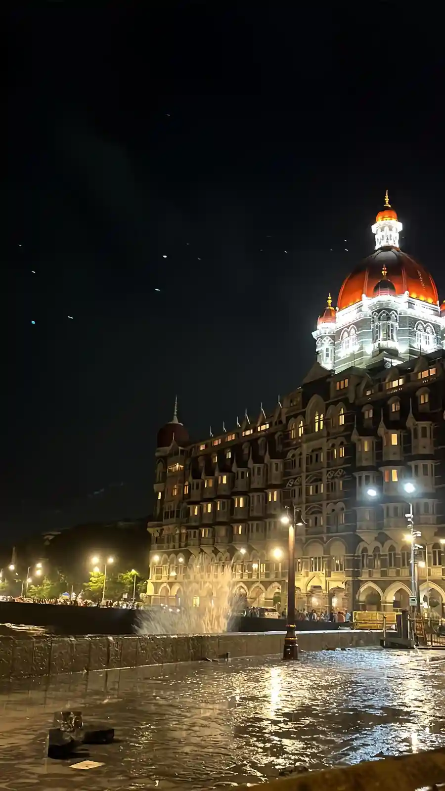 A nighttime photo shows the iconic Taj Mahal Palace Hotel in Mumbai, its grand domes and arches illuminated against a dark…