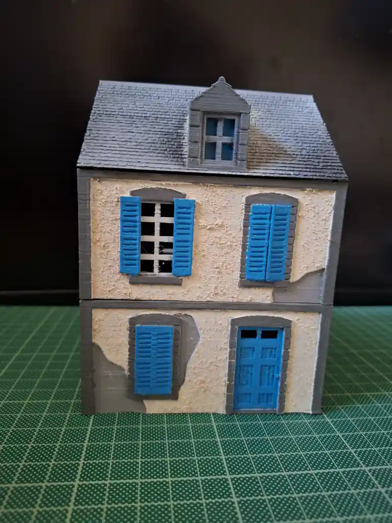 A 3D-printed miniature house model with beige walls, grey trim, and bright blue shutters and door, sitting on a green grid…