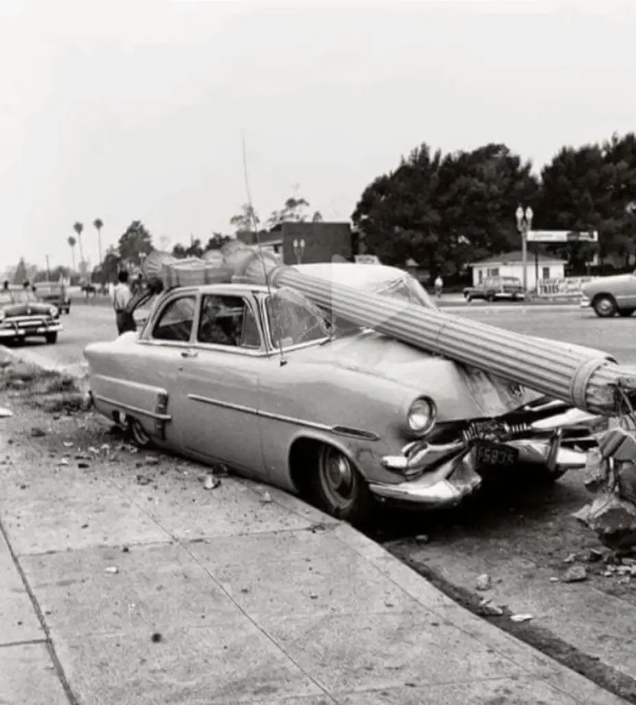 A black-and-white photo shows a vintage 1950s car parked on a street curb, with a large, thick pole or pipe resting…
