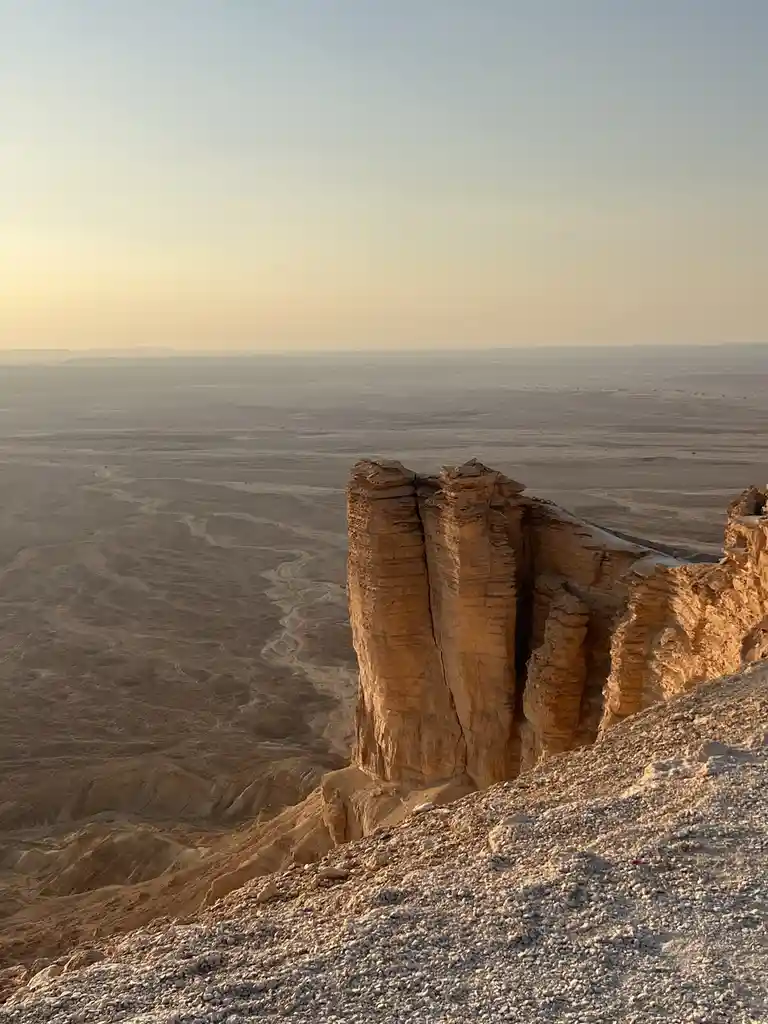 A wide-angle photo captures a vast desert landscape at sunset, with towering, layered sandstone cliffs in the foreground…