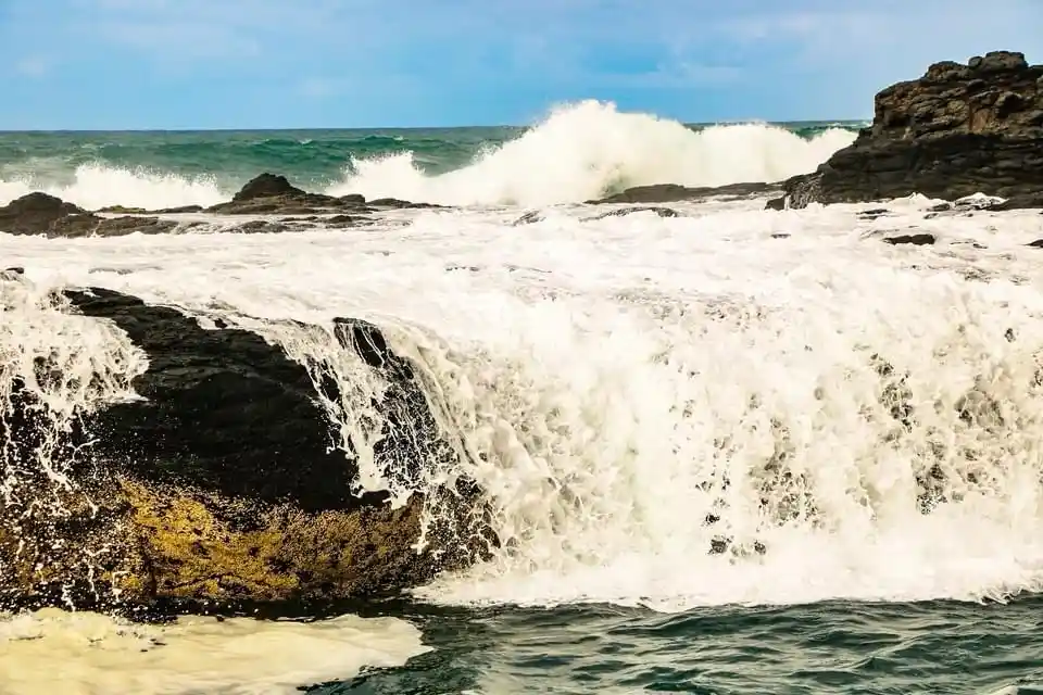 A dynamic ocean scene shows powerful waves crashing against dark, jagged rocks, creating white foam and spray. The water…