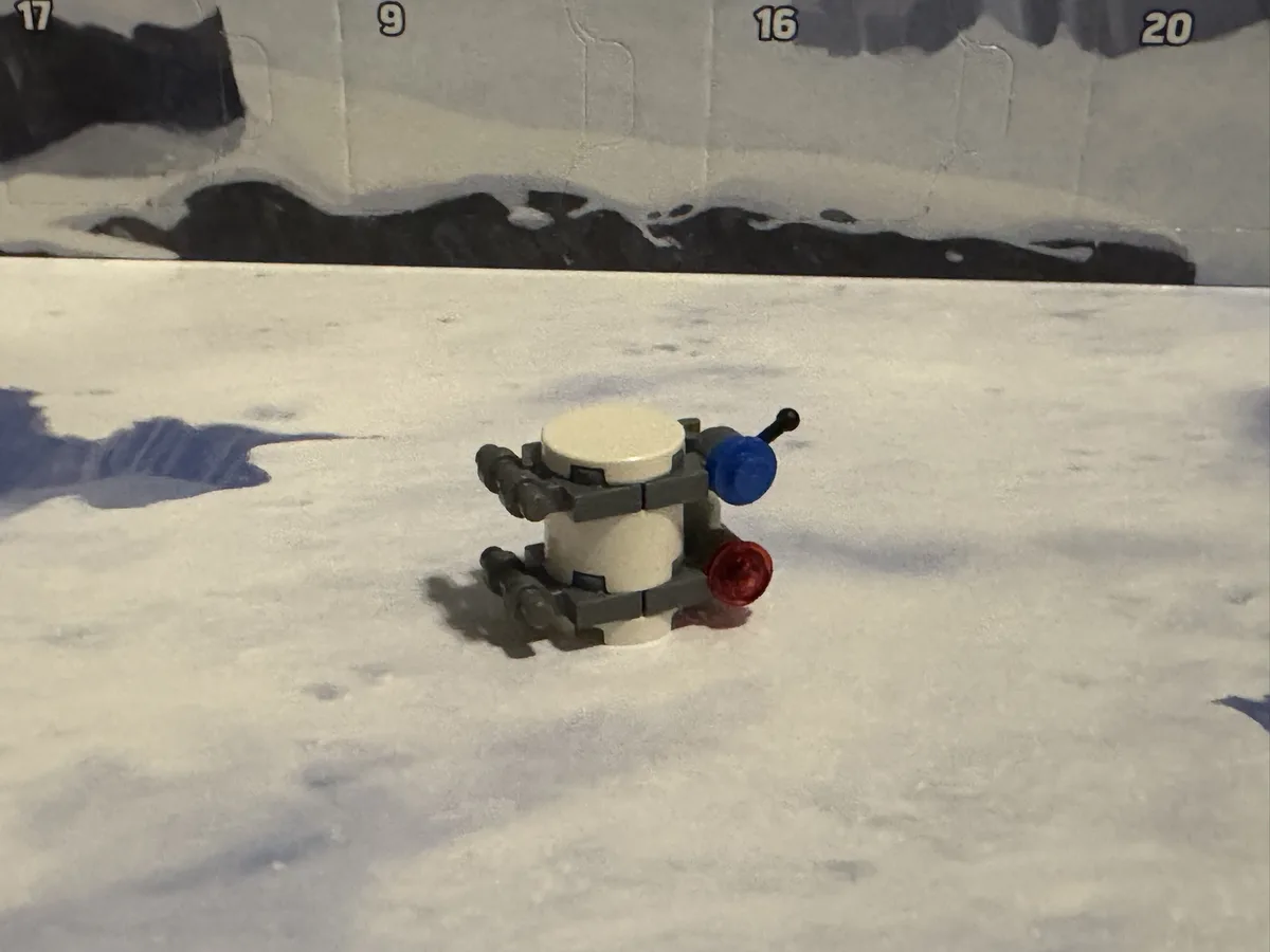 A small, crudely assembled LEGO model resembling a robot or vehicle sits on a snowy surface, with a backdrop showing…