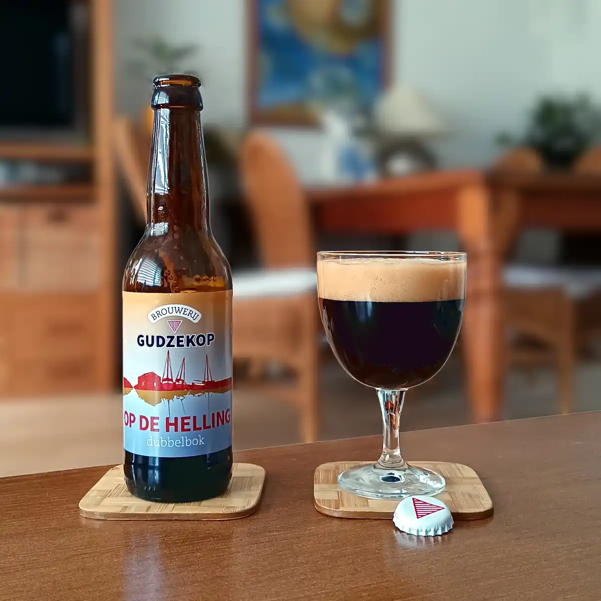 A brown beer bottle labeled 'Brouwerij Gudzekop Op de Helling dubbelbok' sits next to a glass of dark beer with a thick tan…