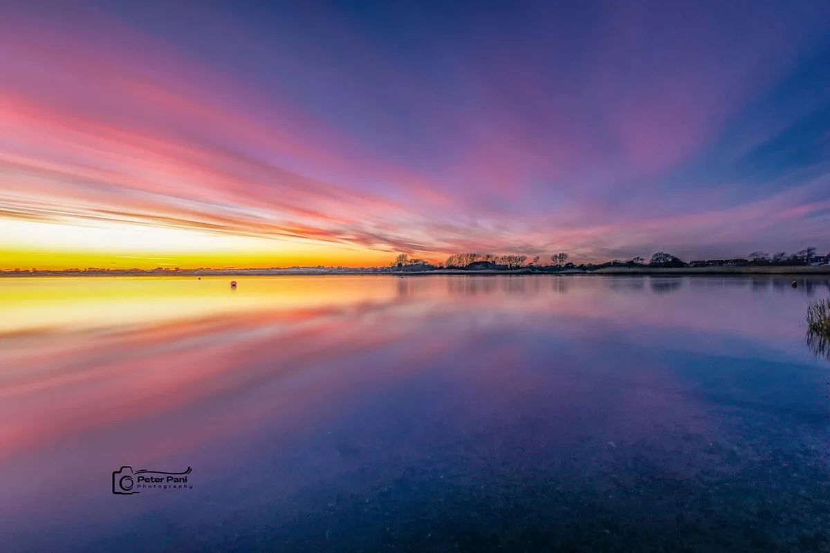 A long-exposure photograph of a serene lake at sunset, with streaks of pink, orange, and purple clouds reflected in the…