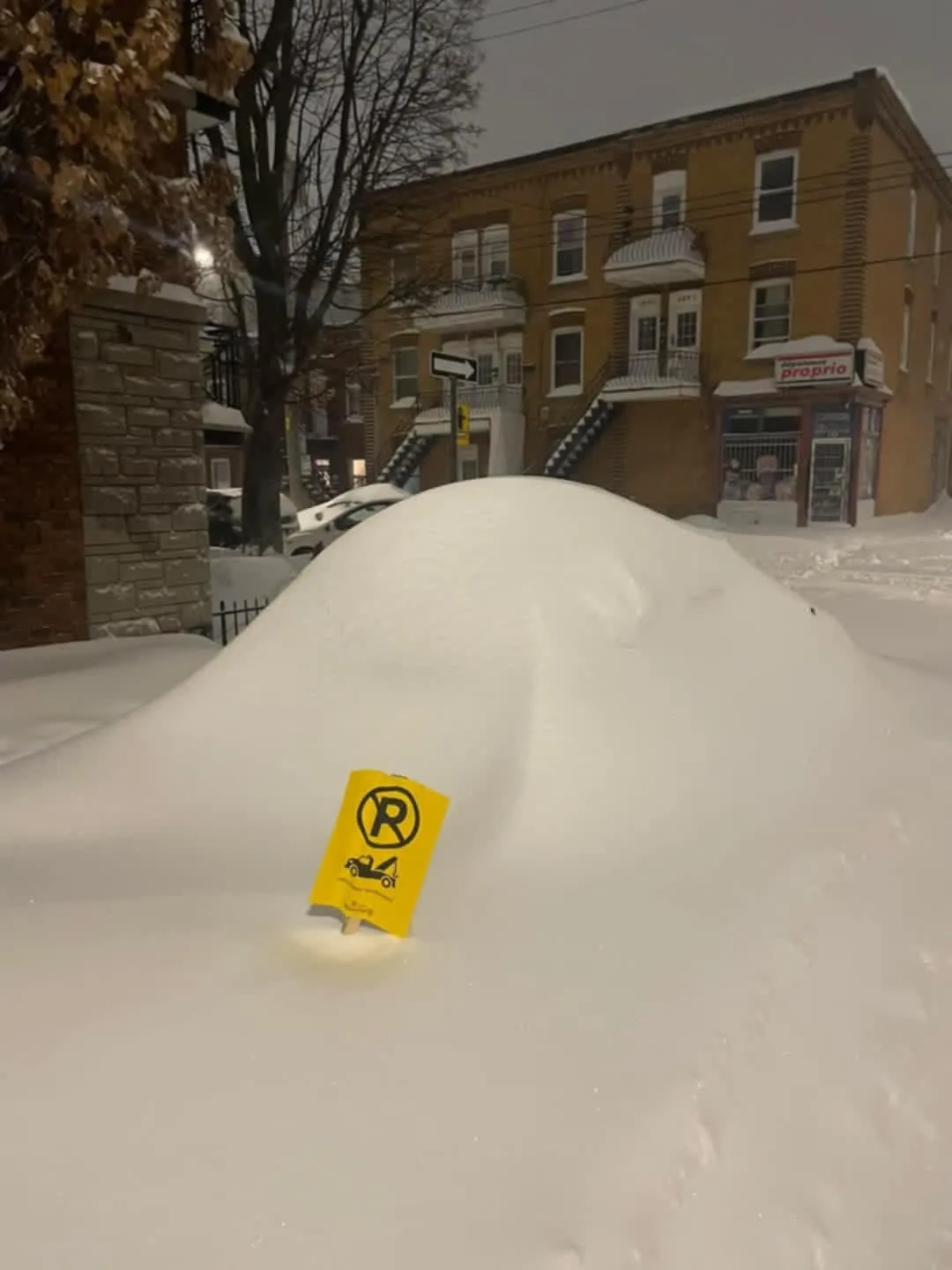 A large mound of snow covers a parked car on a snowy street, with a yellow 'no parking' sign stuck in the snow in front of…