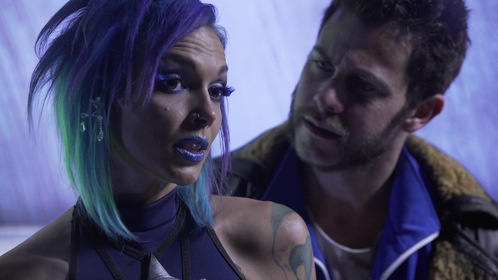A still from a movie or TV show featuring a woman with vibrant purple and green hair, blue lipstick, and tattoos, looking…