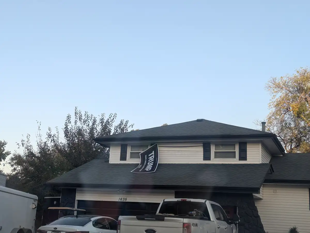 A residential house with a dark roof and white siding has a large, black, curved political banner attached to its side…