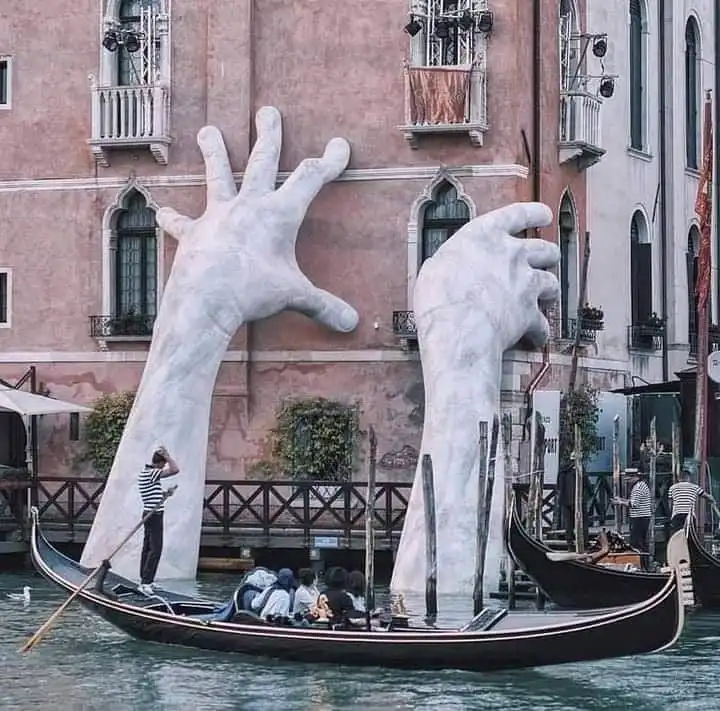 A gondola with passengers and a gondolier floats on a canal in Venice, passing two giant white marble hands emerging from a…