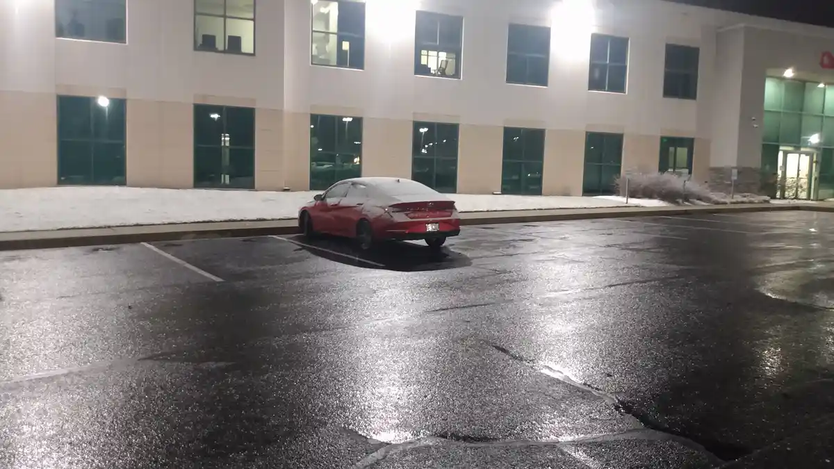 A red sedan is parked in a wet, empty parking lot at night, partially covered in snow, with a lit-up office building behind…
