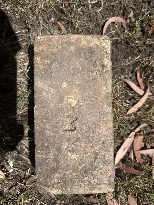 A weathered, rectangular brick lies on grass and dirt, with a faint, stylized 'D' and a squiggly line below it, resembling…