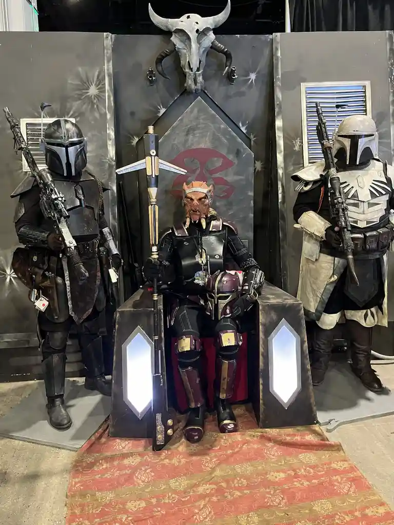 Three people in Mandalorian armor stand guard on either side of a central figure seated on a throne. The central figure…