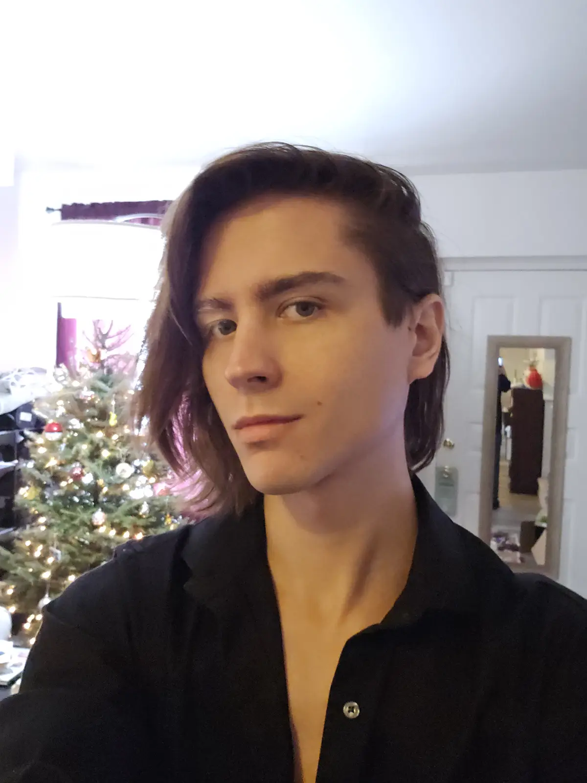 A young man with shoulder-length brown hair and a black button-up shirt looks directly at the camera with a neutral…
