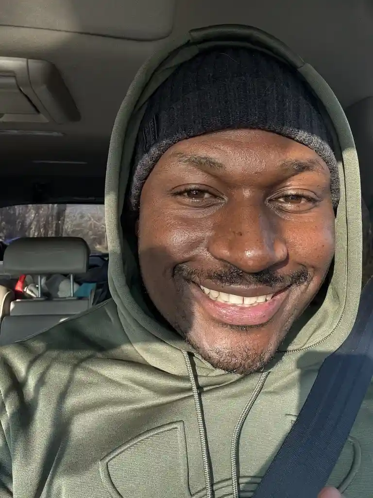A man wearing a green hoodie with the hood up and a black beanie smiles broadly while seated in a car, with a seatbelt…