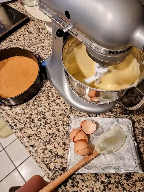 A silver KitchenAid Artisan stand mixer blends a pale yellow batter in its bowl, with three cracked eggshells visible…