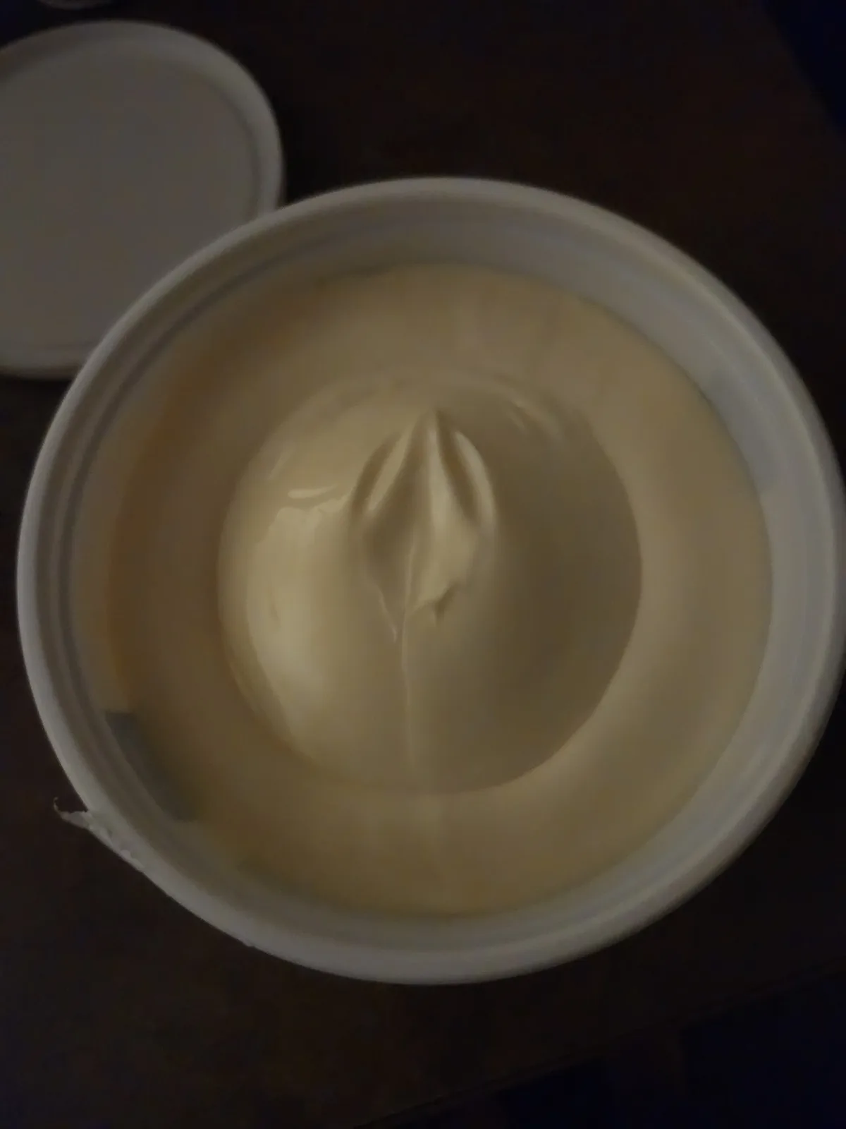 A top-down view of a white plastic tub filled with creamy, pale yellow substance, likely mayonnaise or a similar condiment…