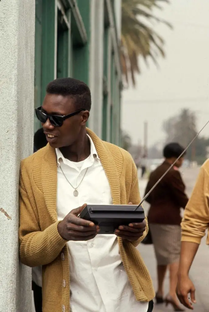 A man in a mustard yellow cardigan and sunglasses leans against a wall while holding a portable radio with an antenna. The…