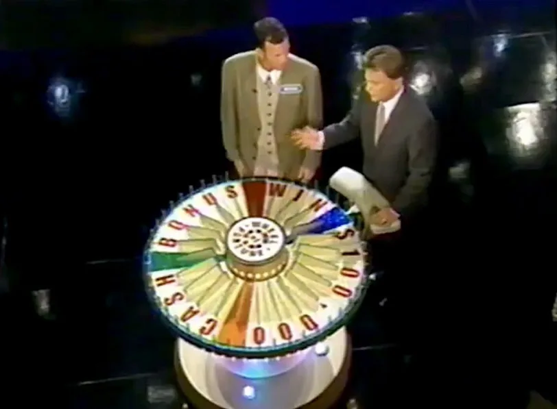 Two men stand beside a large, colorful game show wheel with sections labeled 'CASH', 'BONUS', 'WIN', and '$100'. The man on…