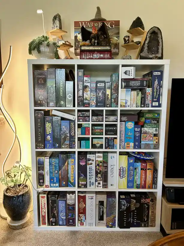 A white bookshelf is densely packed with hundreds of board game boxes, arranged neatly in rows and columns. On top, there…