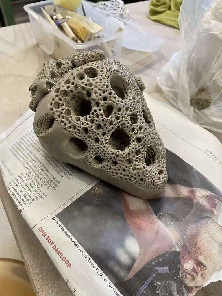 A clay sculpture of a human skull with numerous holes and textured details rests on a newspaper featuring a photo of a…