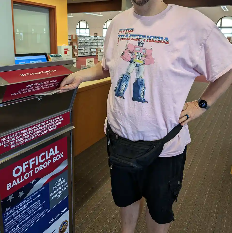 A person wearing a pink and blue Optimus Prime t-shirt with the text 'STOP TRANSPHOBIA' is inserting a ballot into an…