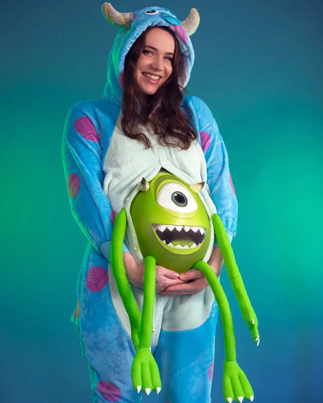 A woman in a blue, polka-dotted Sulley onesie from Monsters, Inc. is smiling while holding a green, one-eyed Mike Wazowski…