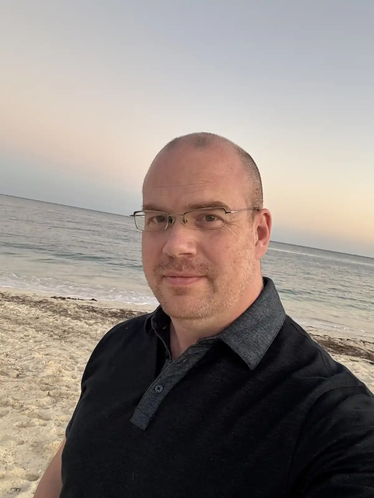 A man with glasses and a shaved head takes a selfie on a sandy beach at sunset, wearing a black polo shirt. The overlaid…