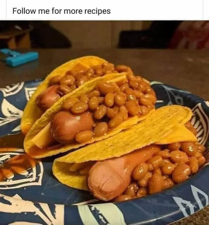 Two hot dogs in yellow corn tortillas, generously topped with baked beans, are served on a blue and white patterned paper…