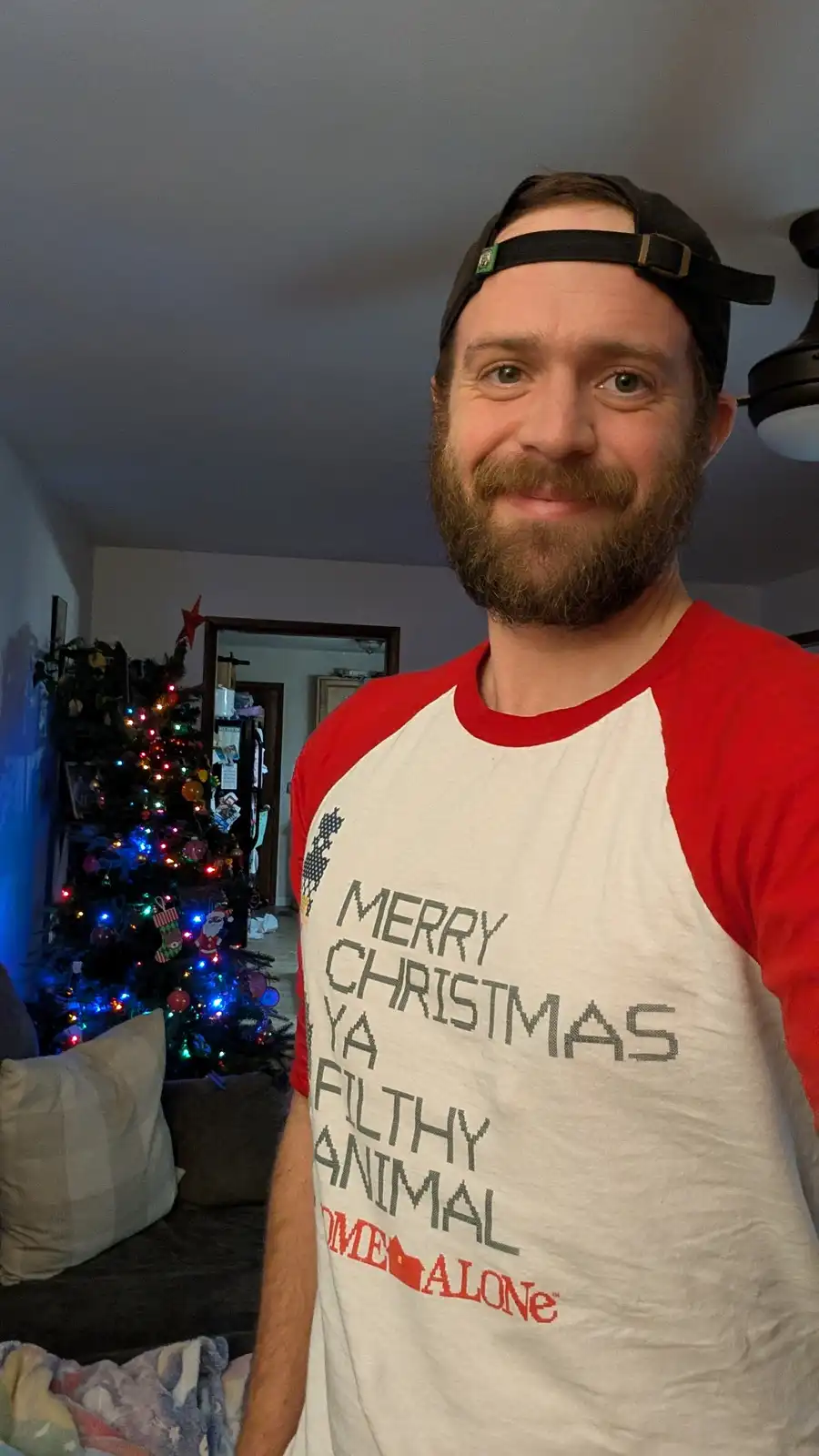 A man with a beard and a black cap wears a red-and-white Christmas-themed t-shirt that reads 'MERRY CHRISTMAS YA FILTHY…