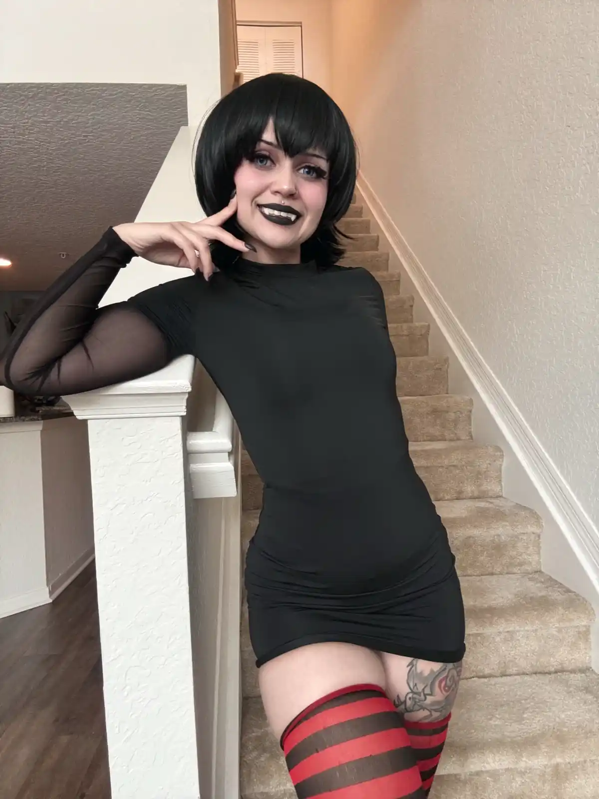 A person with short black hair and black lipstick poses on a staircase, wearing a black dress, red-and-black striped…