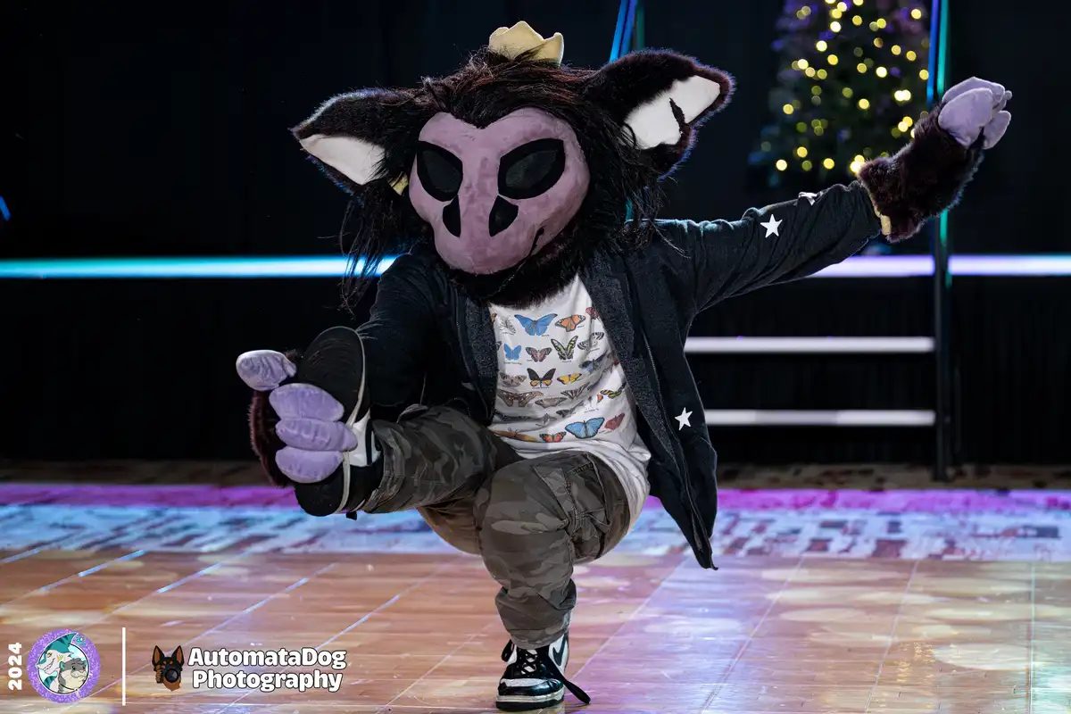 A person in a furry costume with a purple, masked face, black fur, and large ears strikes a dynamic dance pose on a stage…