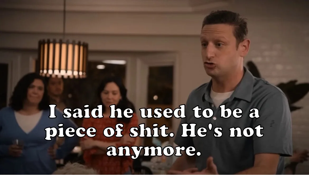 A man in a grey polo shirt is speaking with a surprised or earnest expression in a social setting, with blurred people and a…
