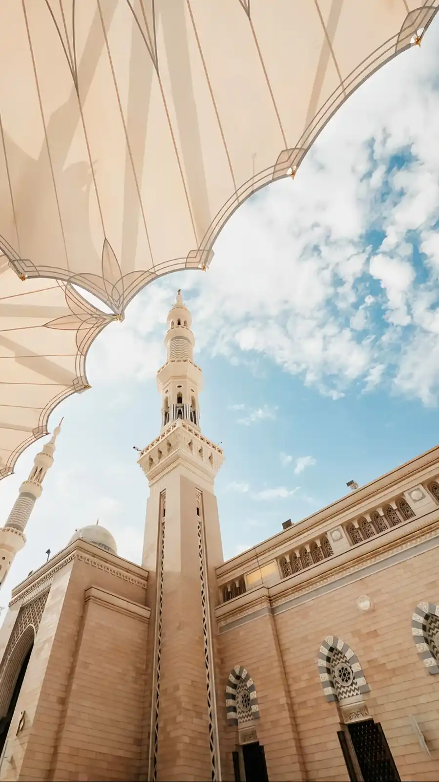 A low-angle photo shows a large beige outdoor umbrella framing a view of a mosque's minaret against a blue sky with clouds…