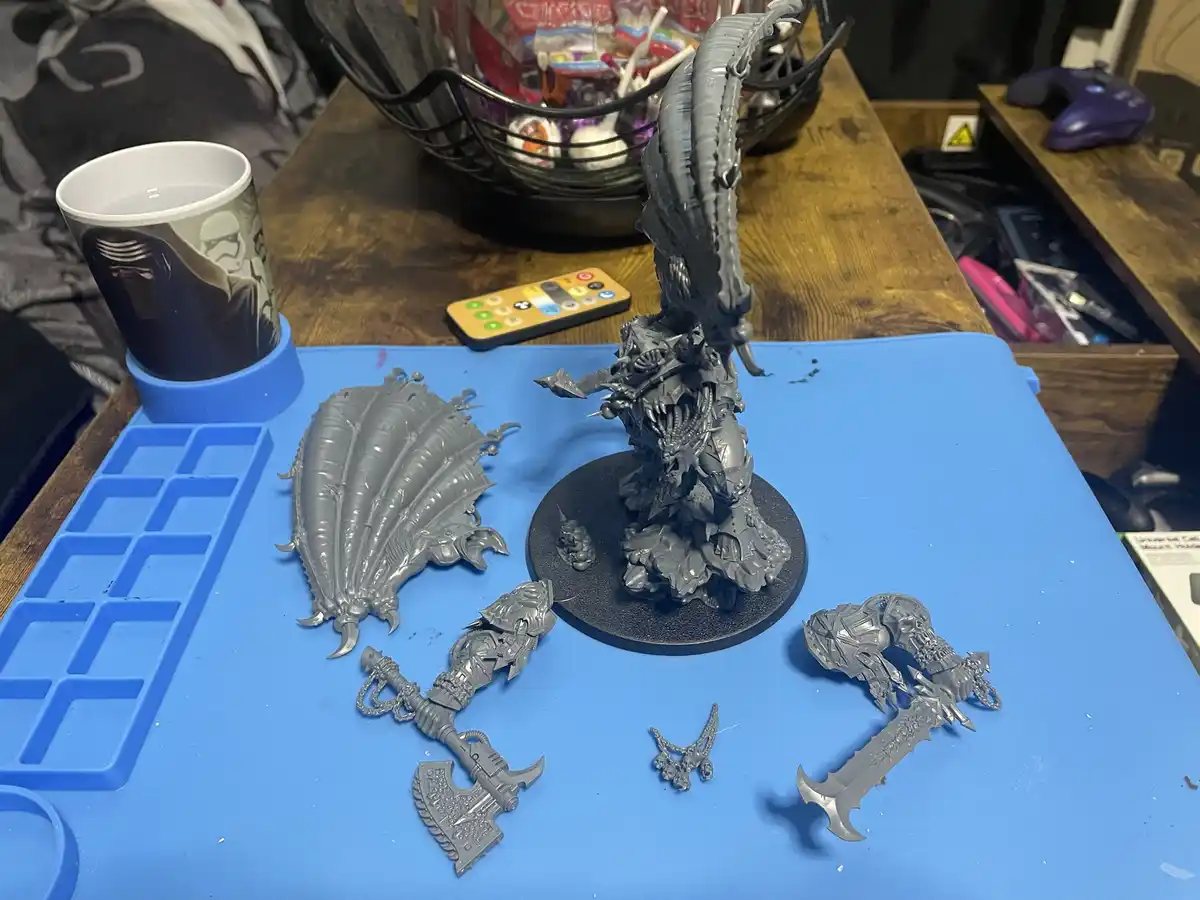 A photo shows a partially assembled grey plastic fantasy miniature on a black base, surrounded by its detached parts like…