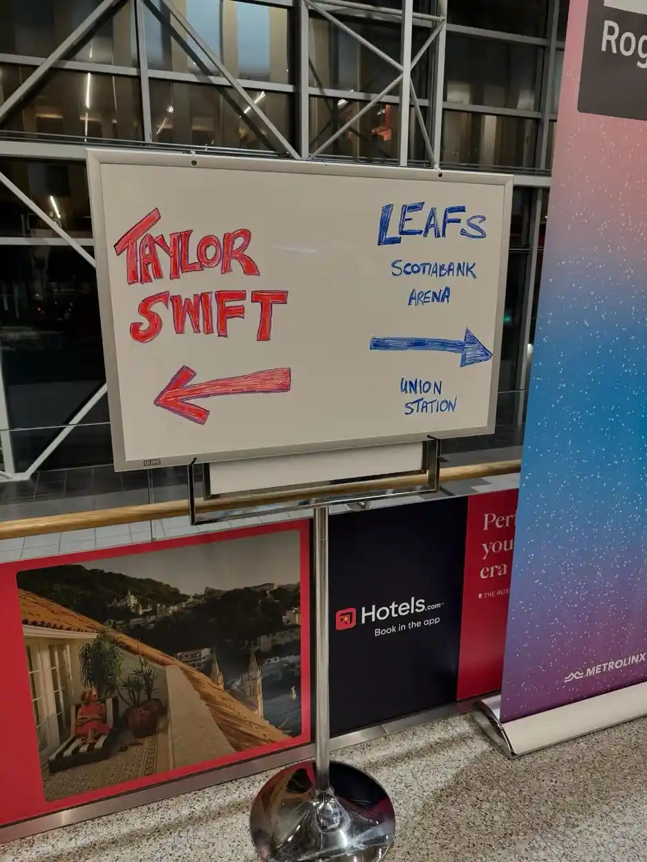 A whiteboard sign in a transit station has 'TAYLOR SWIFT' written in red with an arrow pointing left, while 'LEAFS…